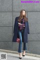 A woman standing in front of a gray wall wearing a blue coat.